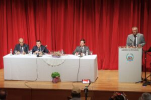 A panel discussion with four men at a table, set against a red curtain backdrop. One man is speaking at a podium.