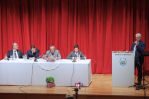 A panel discussion on stage with five men seated at a table, a speaker at a podium, and a red curtain backdrop.