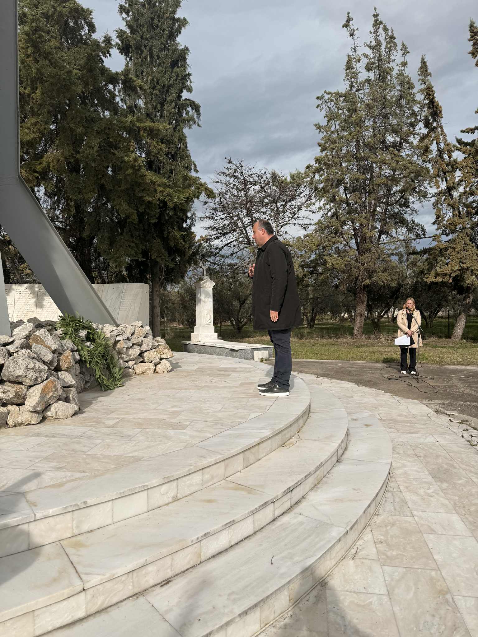 A man stands on marble steps, speaking into a microphone, with trees and a statue in the background. Another person is nearby, holding papers.