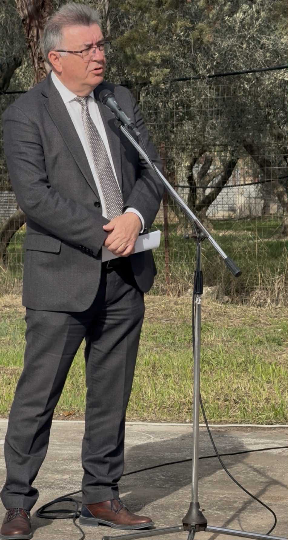 A man in a suit stands at a microphone outdoors, speaking with a serious expression. Green grass and trees are in the background.