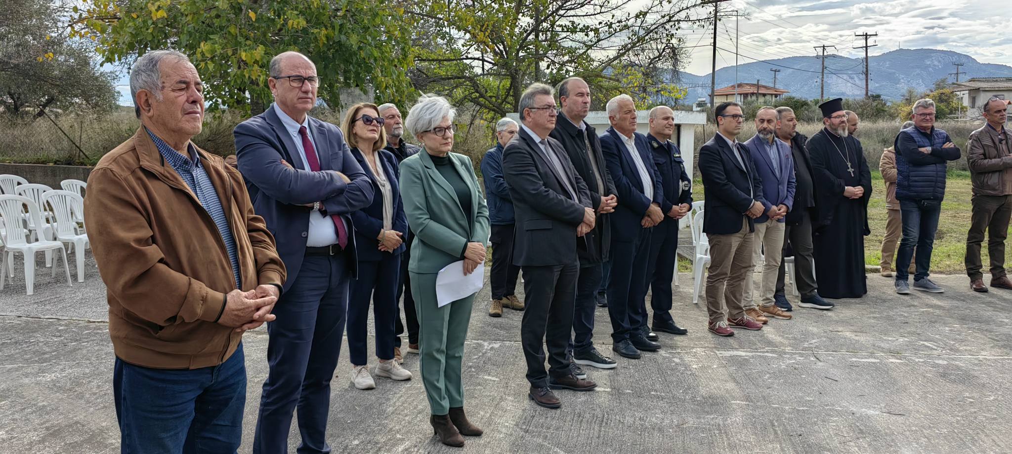 A group of people stands in a line outdoors, some in formal attire, with trees and empty chairs in the background.