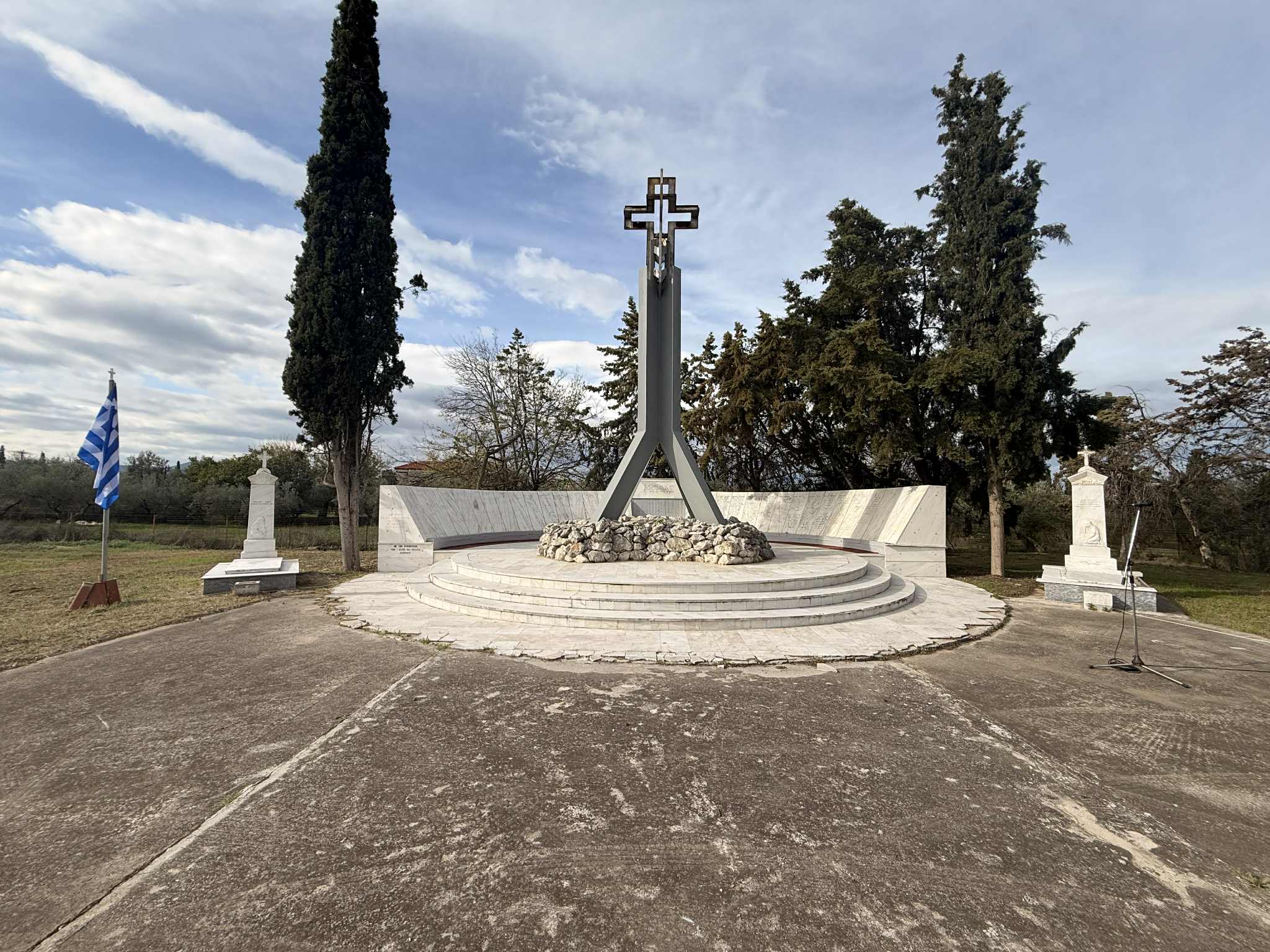 A memorial site featuring a large cross, surrounded by stone and marble structures, with a Greek flag nearby and trees in the background.