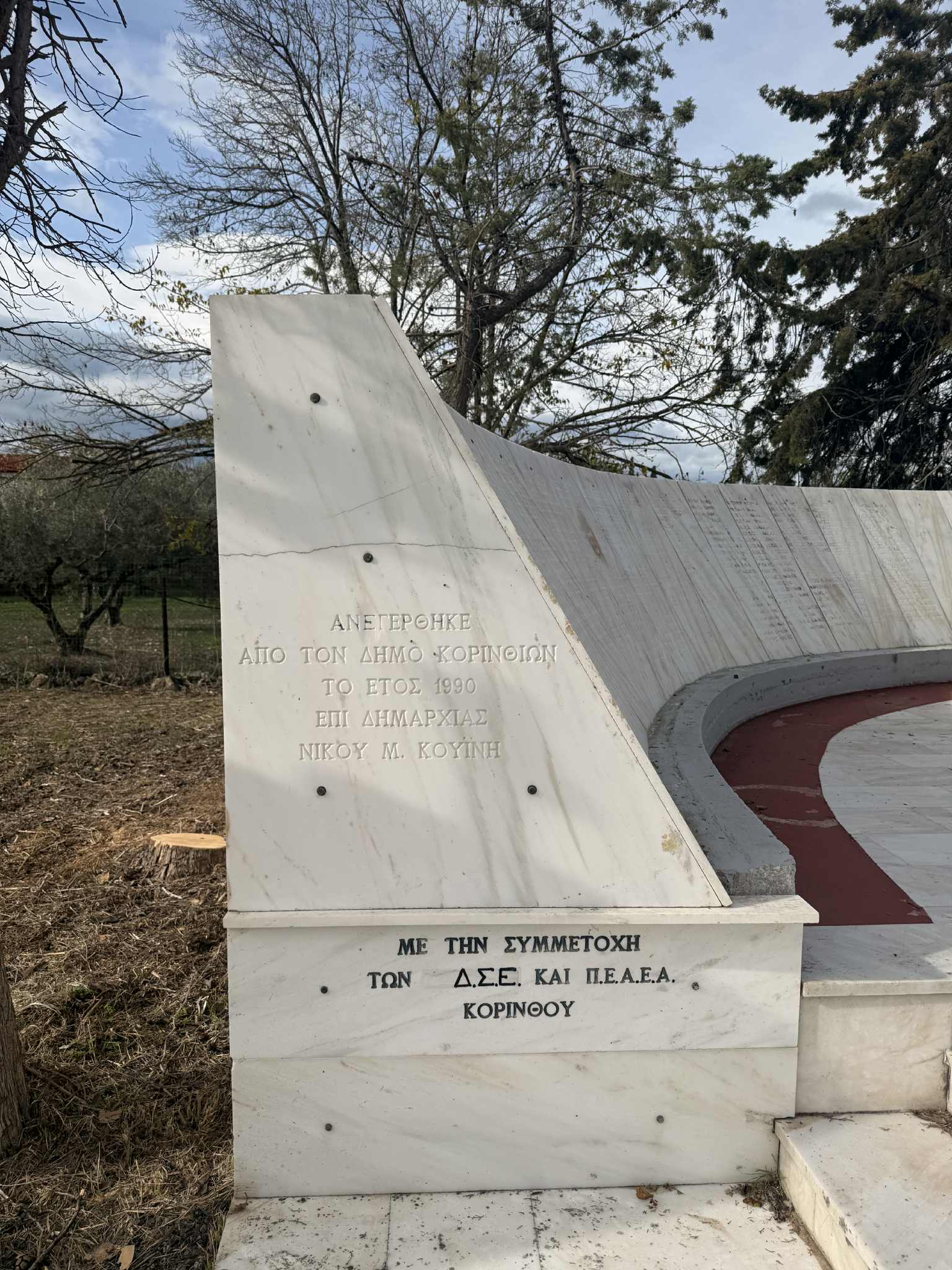 A marble monument with inscriptions, surrounded by trees and a pathway, commemorating an event from 1990 in Korinthos.