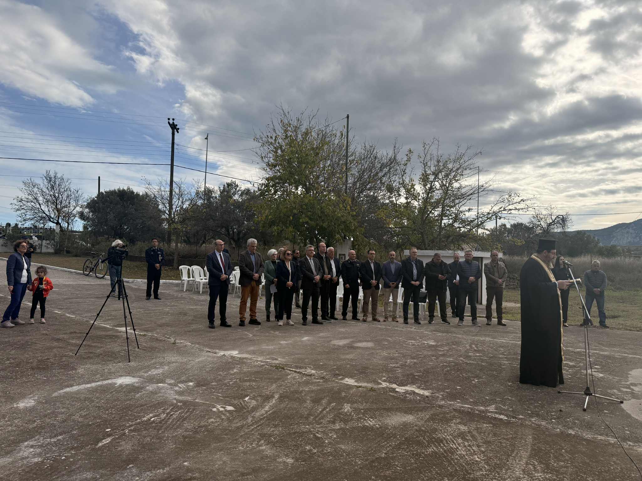 A group of people, including a speaker in traditional attire, gather outdoors for an event under a cloudy sky.