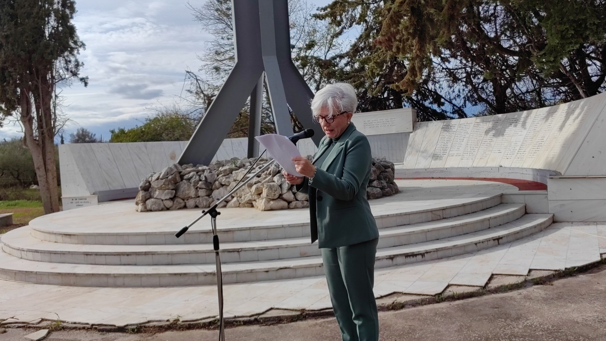 A woman in a green suit reads from a paper at a memorial site with a stone structure and inscriptions in the background.