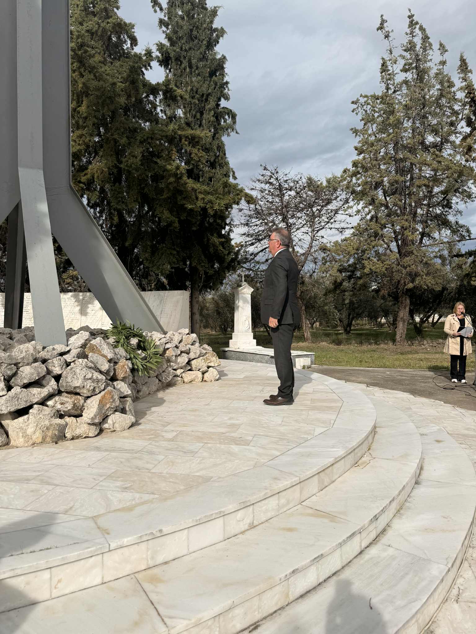 A man in a suit stands on marble steps, gazing at a monument surrounded by trees. Another person is visible in the background.