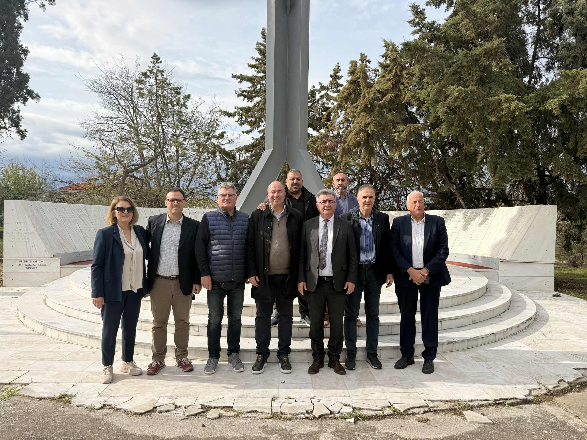 A group of people stands together in front of a monument, surrounded by trees and a cloudy sky.