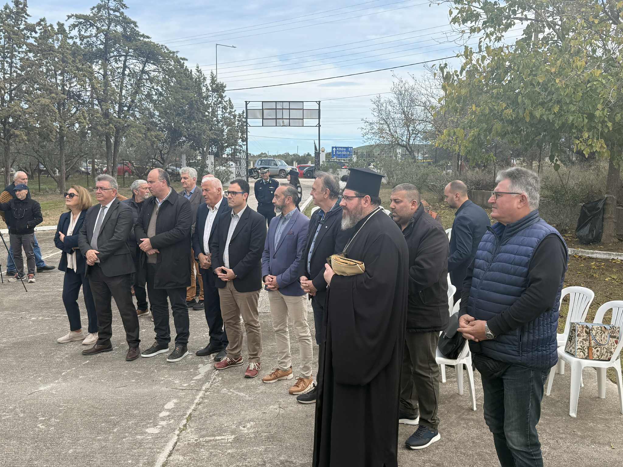 A group of people, including a clergy member, stands together outdoors, with trees and vehicles in the background.