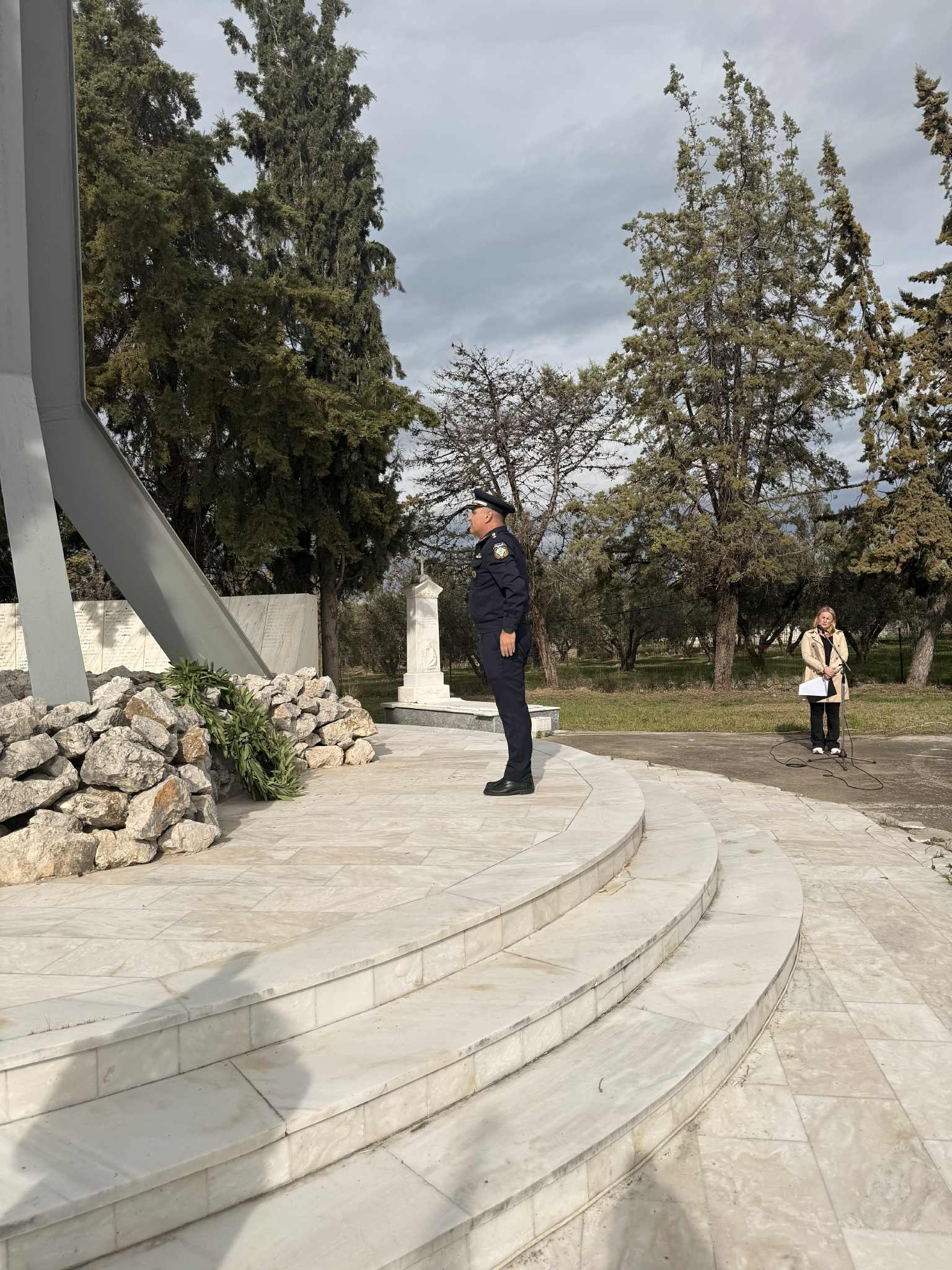 A uniformed officer stands on marble steps near a memorial, surrounded by trees, while a woman with a microphone observes.